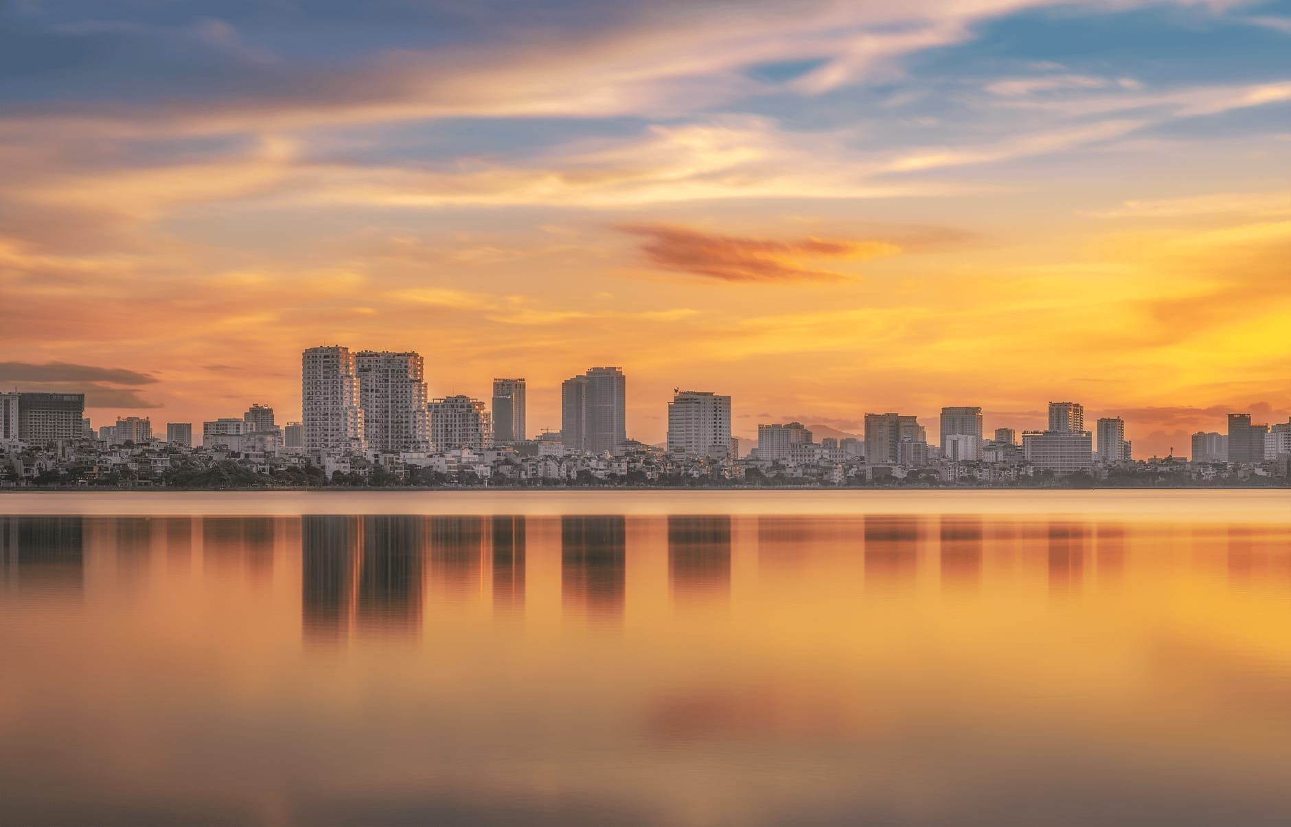 Wide panoramic view of West Lake in Hanoi at sunset, with modern buildings and colorful sky reflecting on calm water.