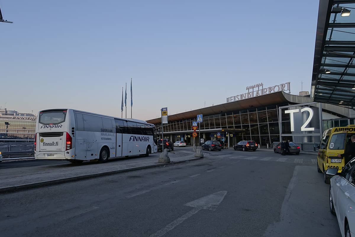 Exterior do Terminal 2 do Aeroporto de Helsinki com sinalização em destaque, ônibus da Finnair, táxis e passageiros do lado de fora durante o dia.