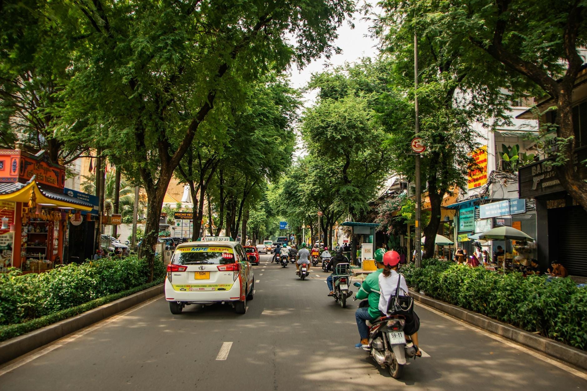Một con phố yên tĩnh, rợp bóng cây ở Thành phố Hồ Chí Minh với xe ô tô, xe máy và các cơ sở kinh doanh địa phương, tạo nên bầu không khí khu phố bình yên.