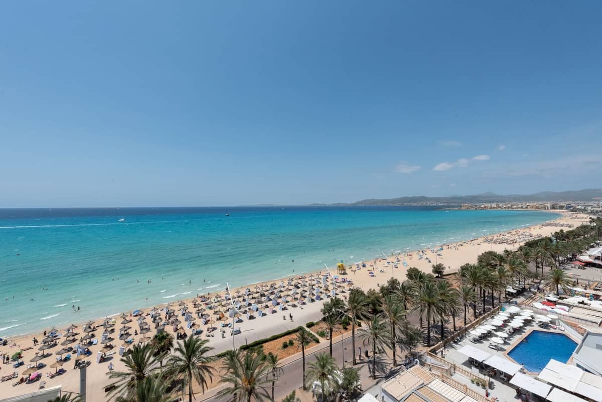 Aerial view of a long sandy beach lined with palm trees, sunbeds, and clear turquoise water next to beachfront hotels.