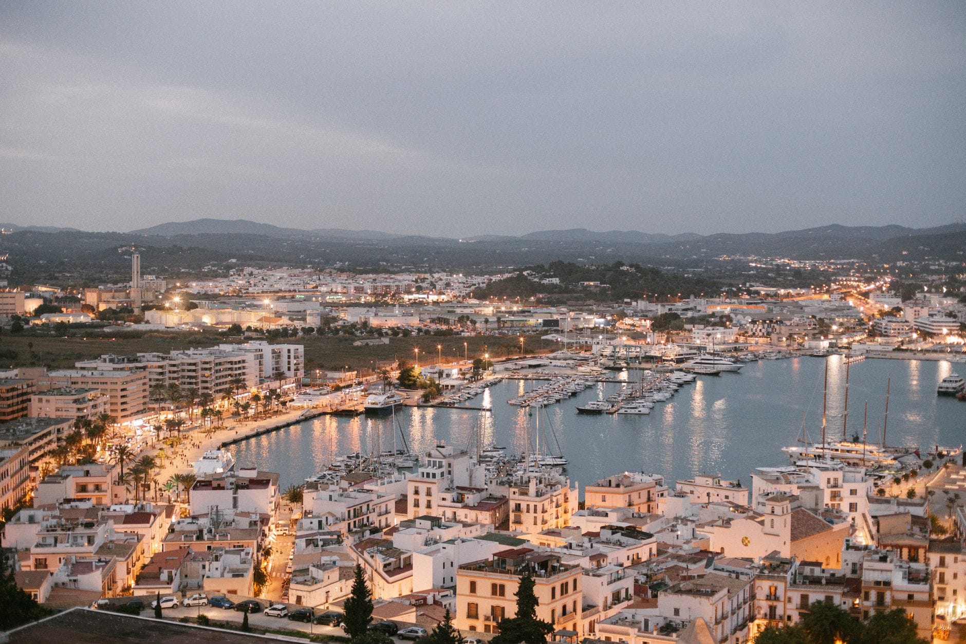 Vista panorâmica ao entardecer da cidade de Ibiza e sua marina, com edifícios brancos e iates enfileirados, colinas ao fundo e as luzes da cidade começando a brilhar.