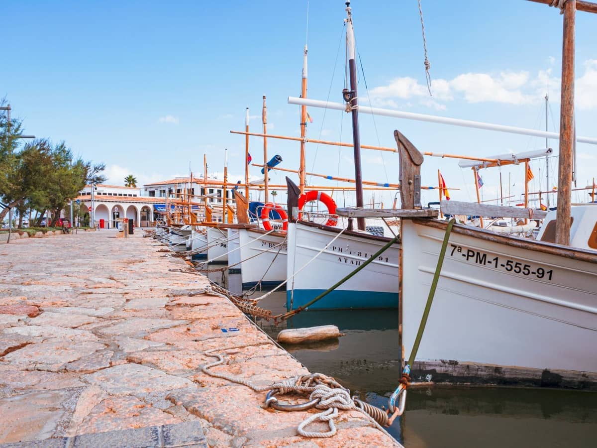 Traditional wooden boats moored along a stone promenade in a quiet marina, with white buildings in the background and a calm, blue sky above.