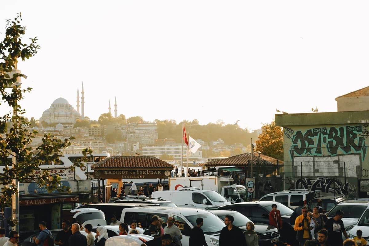 Bulliciosa escena callejera en el lado asiático de Estambul con multitudes, coches, puestos de mercado, cúpulas de mezquitas y cartel de ferry para Kadıköy y Üsküdar.