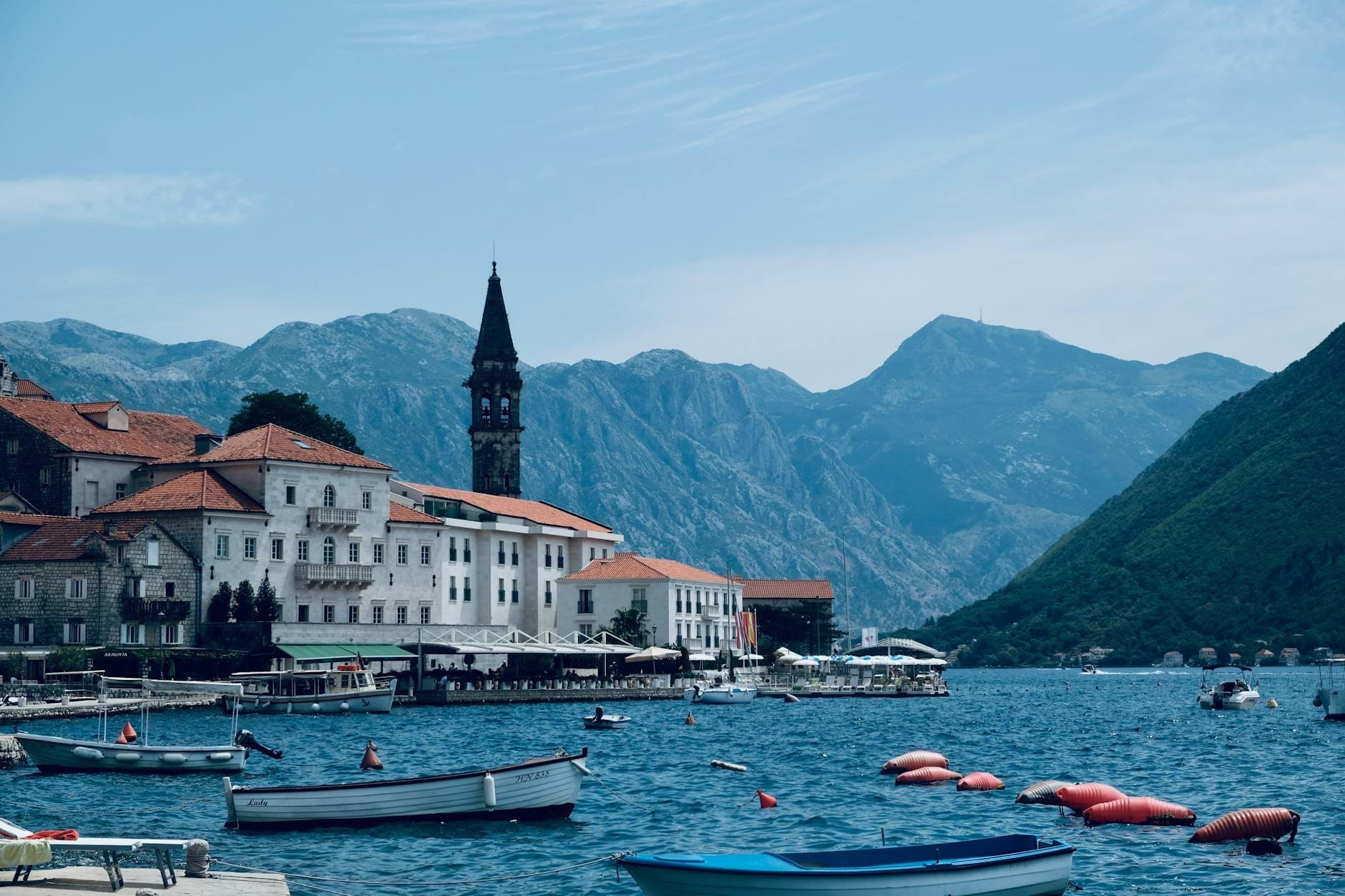 Edifícios medievais de pedra com telhados vermelhos alinham-se à beira-mar em Kotor, Montenegro, com barcos flutuando na baía e montanhas imponentes ao fundo.