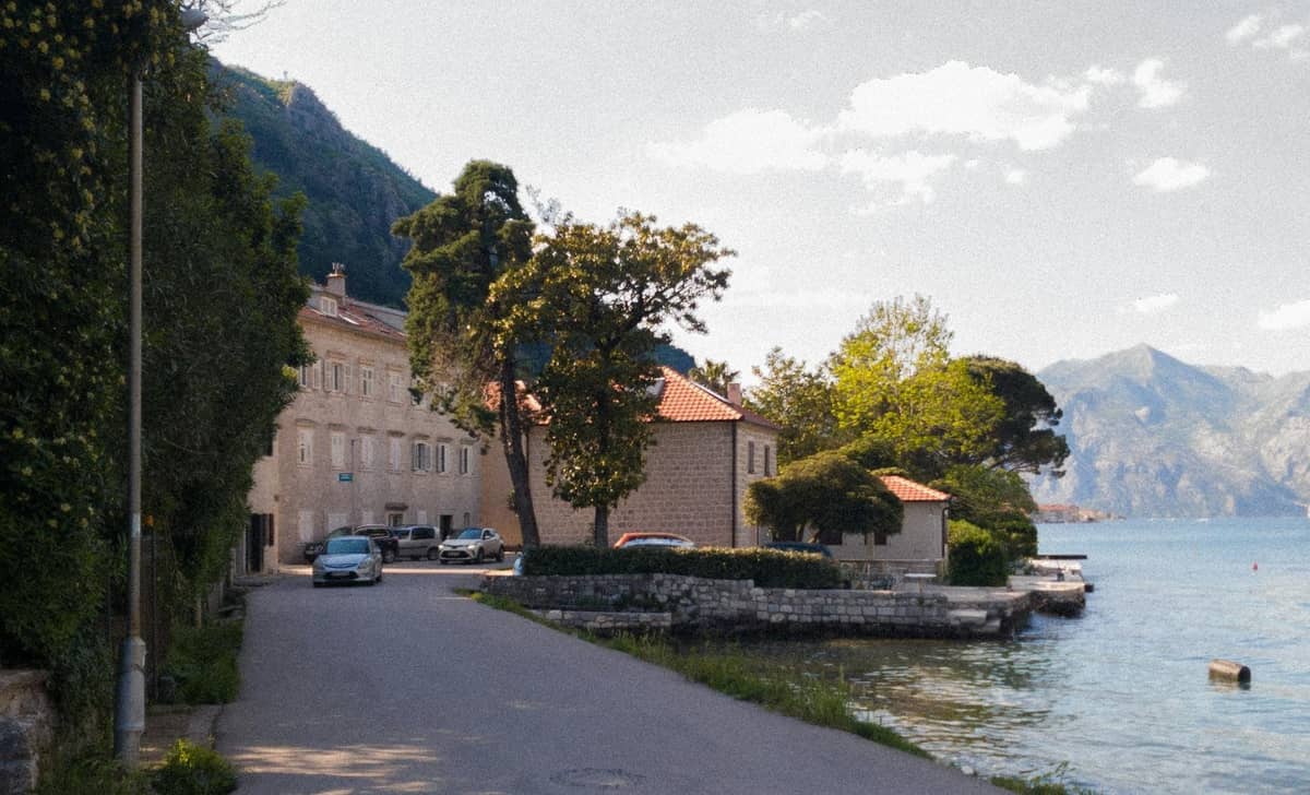 Quaint bayfront road in a quiet village near Kotor, with stone houses, mountains, and calm water.