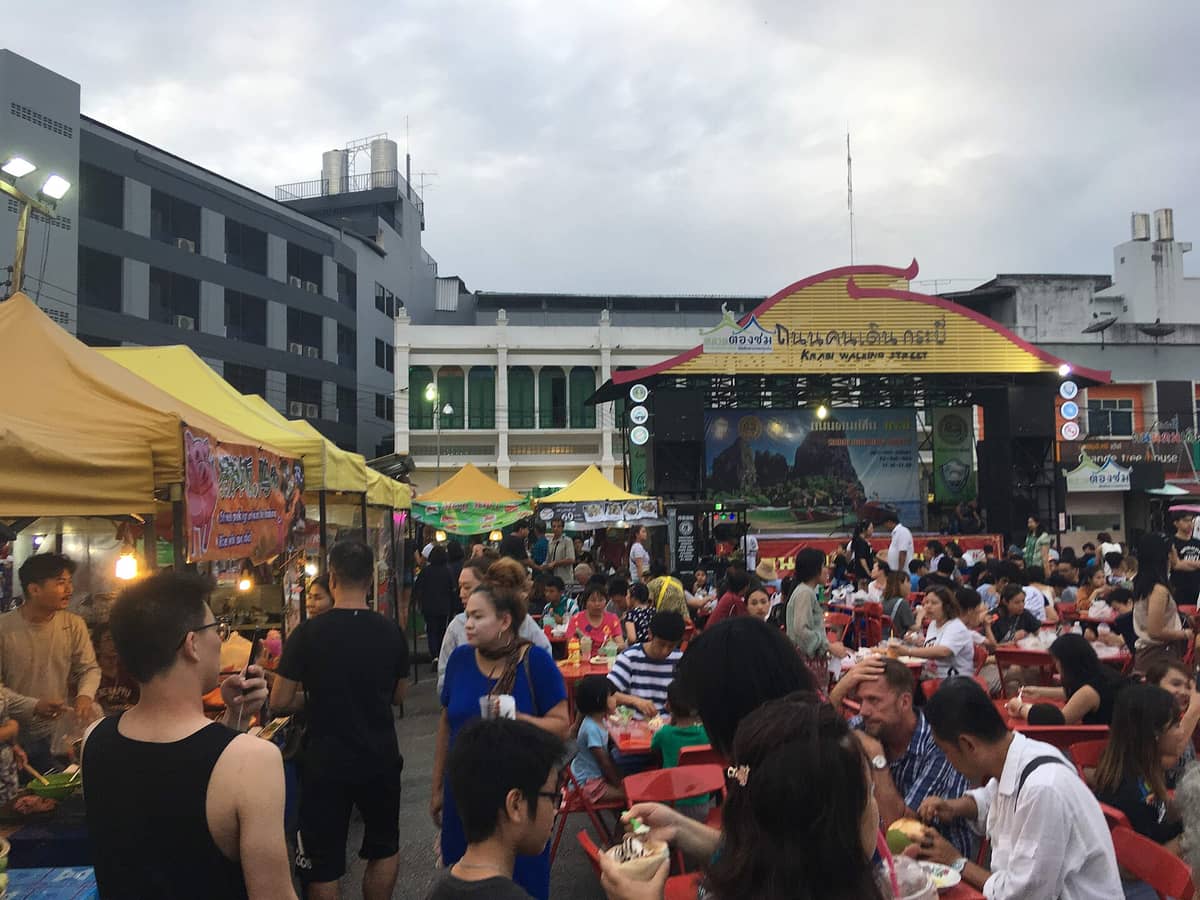 Scène animée du marché nocturne de Krabi Town avec des stands de nourriture sous des tentes jaunes et des foules dînant sur des chaises rouges, une scène portant l'inscription 'Krabi Walking Street' en arrière-plan.