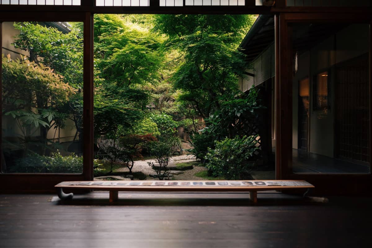 Traditional Japanese ryokan lounge area with wooden floor and large windows overlooking a lush green garden, showcasing serene accommodation style.