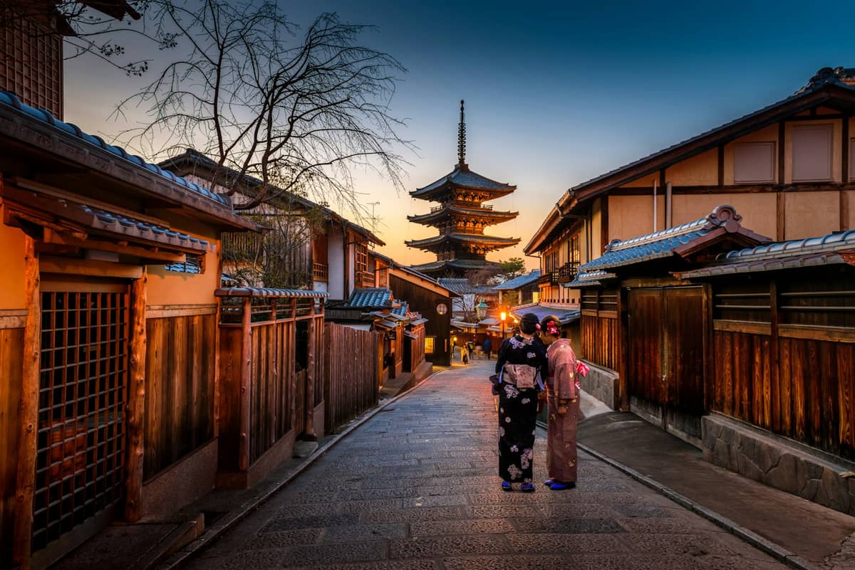 Traditional wooden street in Kyoto at dusk with two people in kimono and the Yasaka Pagoda rising in the background.