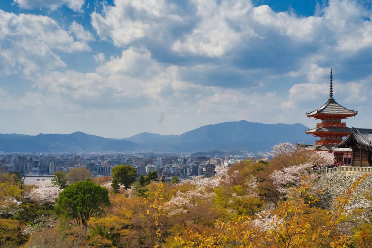 Wide view of Kyoto city with Kiyomizu-dera temple and mountains in the background under a partly cloudy sky.