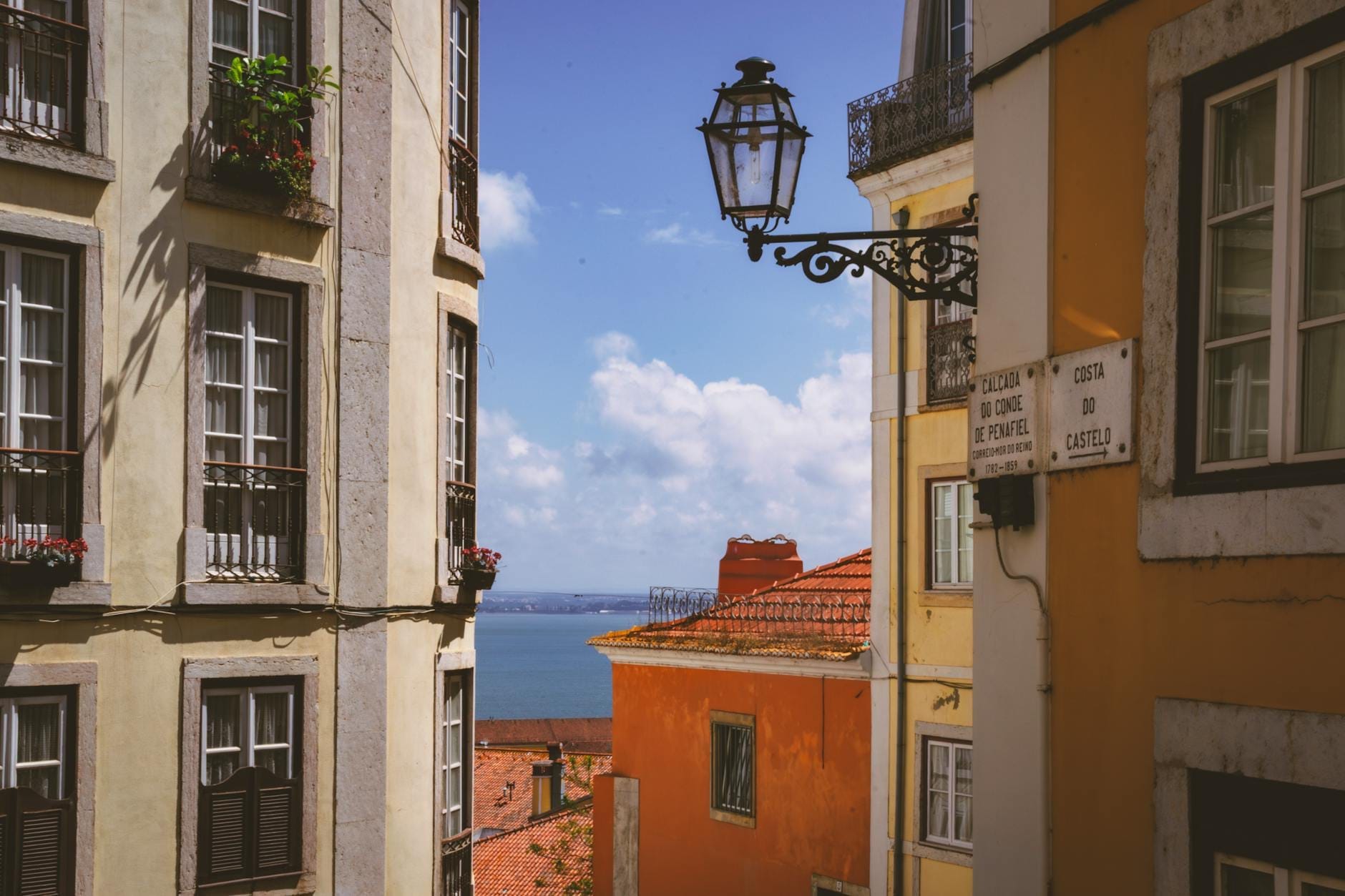 Vista entre os coloridos edifícios de Lisboa com uma luminária decorativa e uma visão do rio Tejo ao longe.