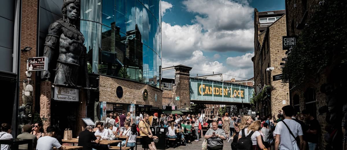 Movimentada área de Camden Lock com multidões, assentos ao ar livre, bancas de mercado e o icônico letreiro da ponte ferroviária em um dia ensolarado.
