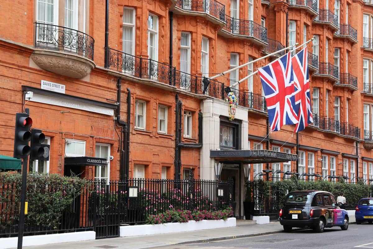Elegante fachada de tijolo vermelho do hotel Claridge's em Mayfair, Londres, com bandeiras Union Jack e um táxi preto londrino do lado de fora.