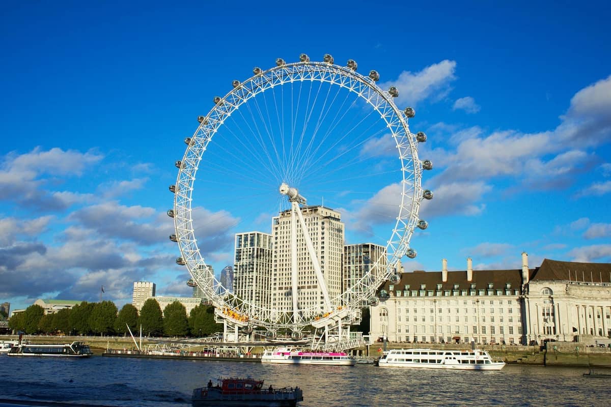 O London Eye na margem sul ao lado do Tâmisa, sob um céu azul brilhante com barcos no rio.