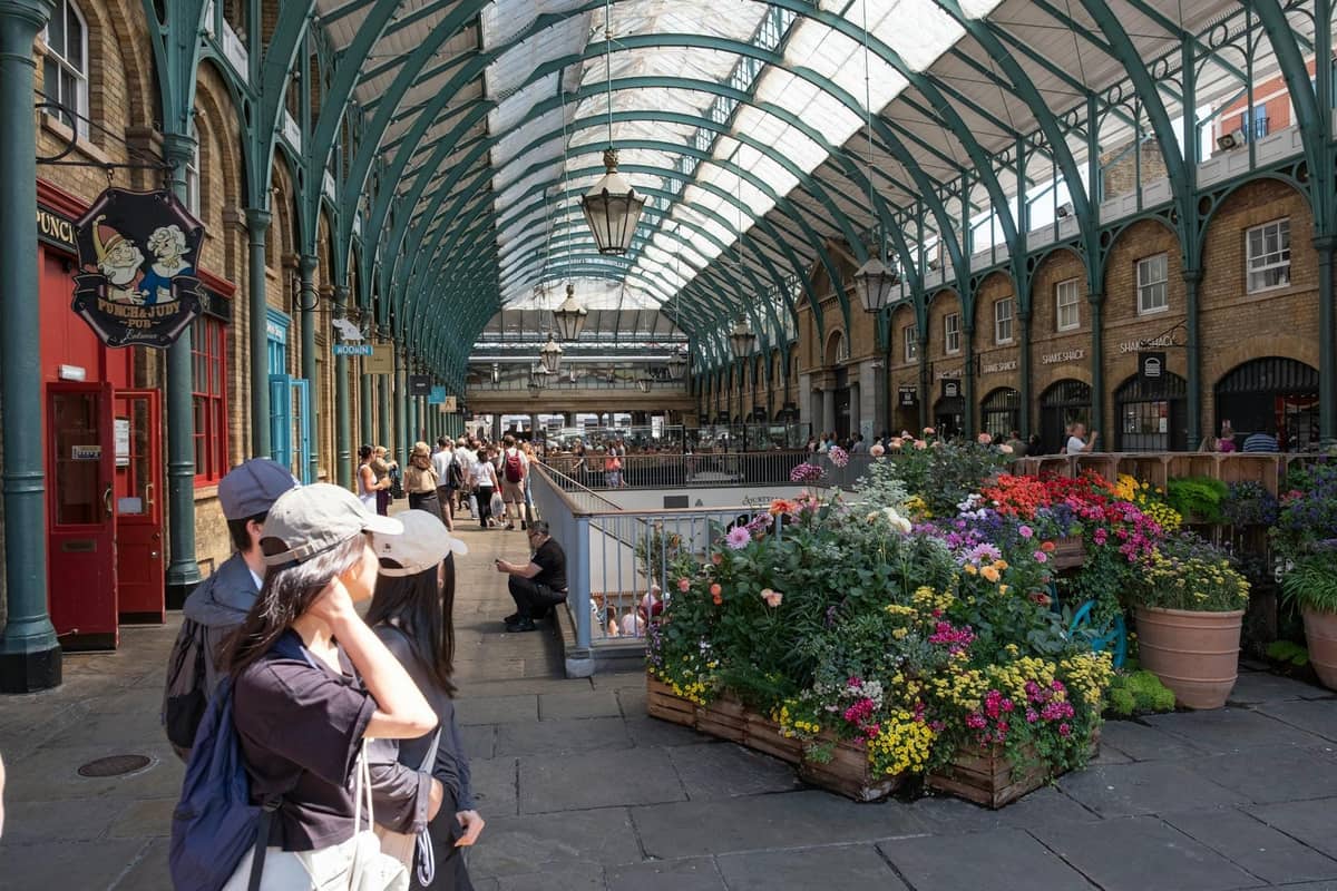 Vista ampla do Covent Garden Market com telhado de vidro, exposições de flores e pessoas explorando o mercado no West End de Londres.