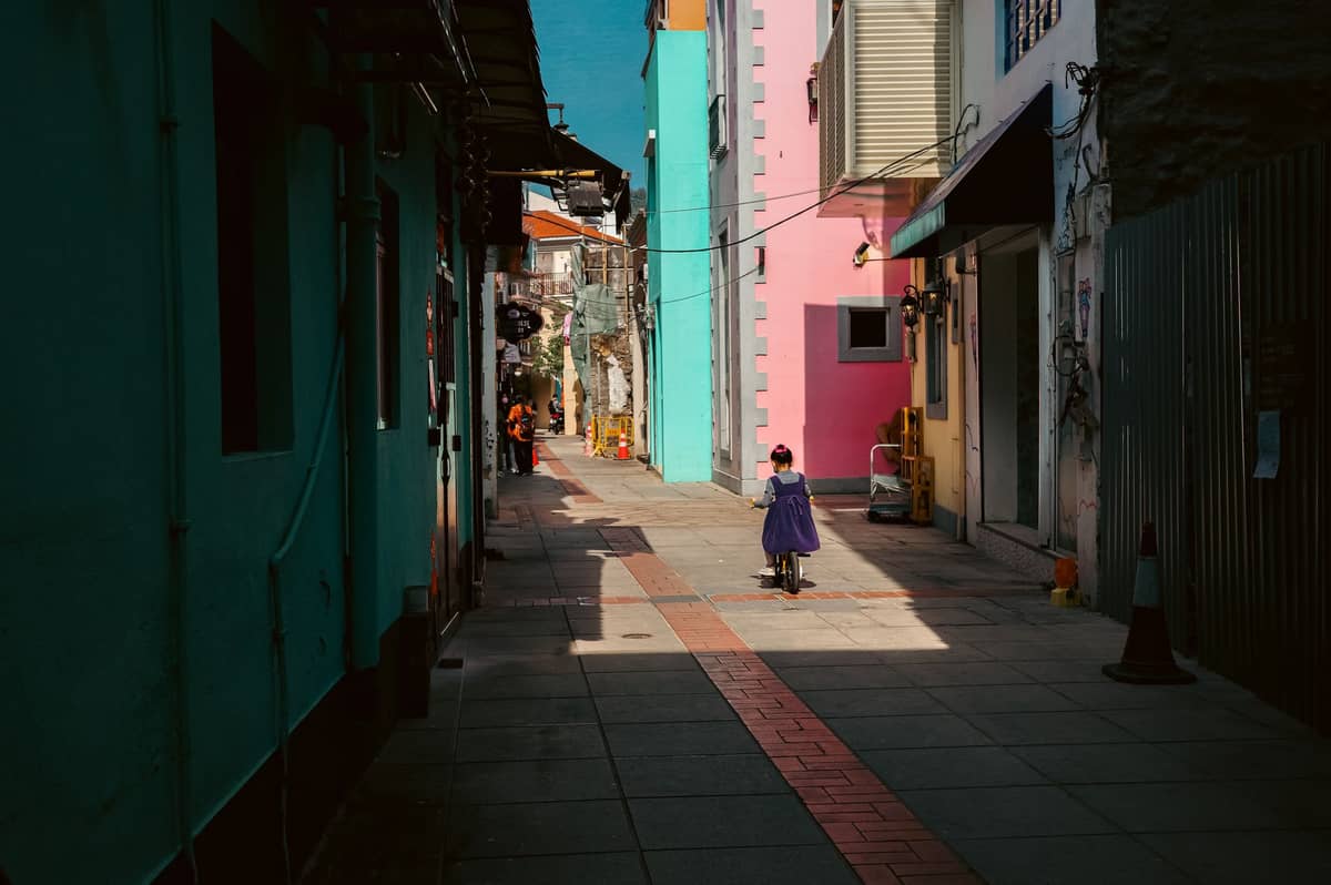 ตรอกแคบในหมู่บ้าน Taipa มาเก๊า พร้อมอาคารสีสันสดใส มีคนเดินอยู่ไกลๆ และแสงแดดยามบ่ายที่ส่องสว่างถนน