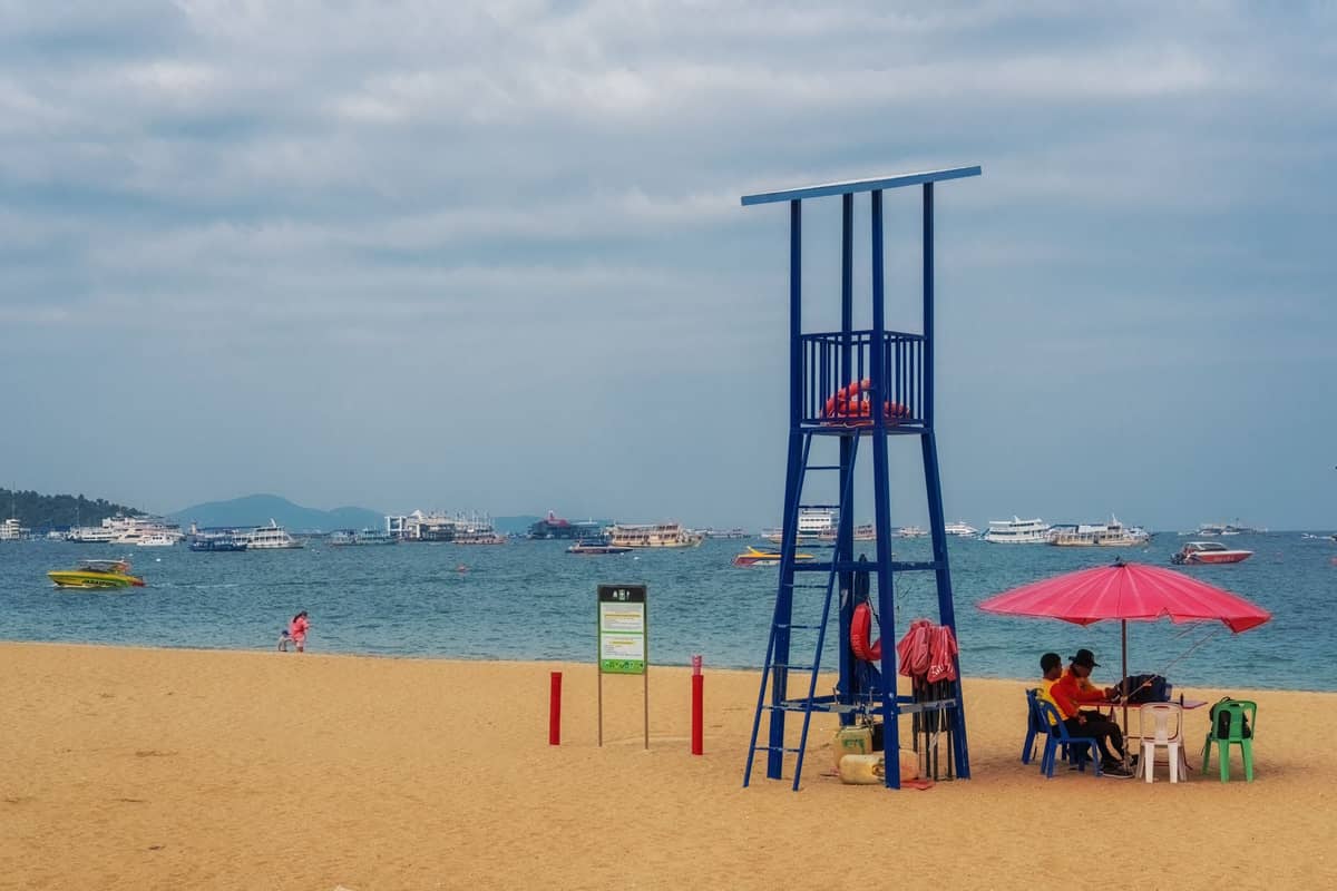 ชายหาดทรายที่มีผู้คนผ่อนคลายใต้ร่มสีชมพูและหอชีวิตรักษา มองเห็นเรือในทะเลภายใต้ท้องฟ้าที่มีเมฆ