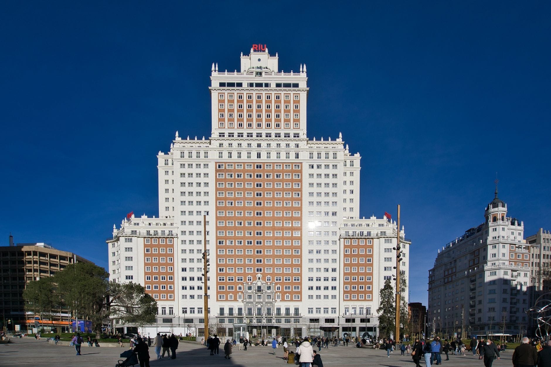 Vista ampla da Plaza de España em Madri com o imponente edifício do hotel ao centro, pessoas caminhando ao redor e um céu azul limpo.