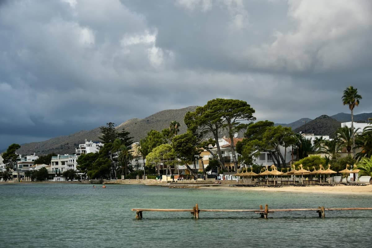Ruhige Strandszene mit klarem Wasser, Sandstrand, Sonnenschirmen und niedrigen Hotels vor grünen Hügeln im Norden von Mallorca.