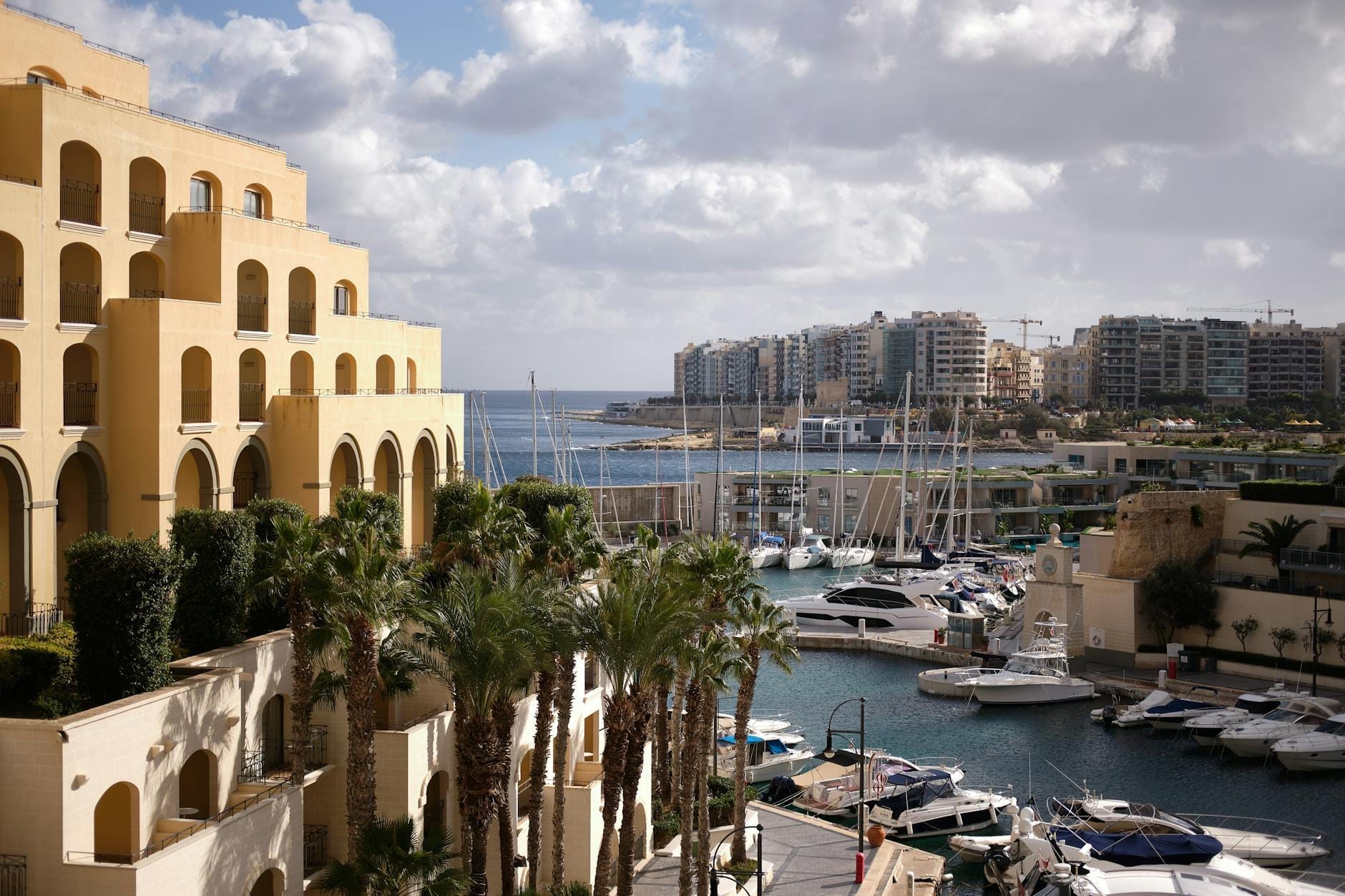 Elegantes edifícios de hotel à beira-mar com palmeiras e uma marina cheia de iates em Malta, com a cidade e o mar ao fundo.