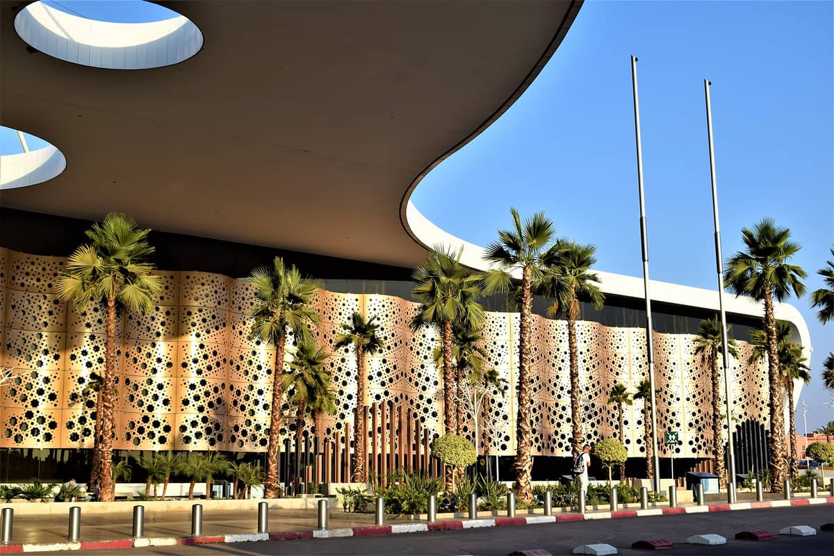 ภายนอกสมัยใหม่ของสนามบิน Marrakech Menara พร้อมด้านหน้าอาคารที่ประดับประดาอย่างวิจิตร ต้นปาล์ม และท้องฟ้าสีฟ้า แสดงถึงประตูสู่การเดินทางเข้าสู่เมือง