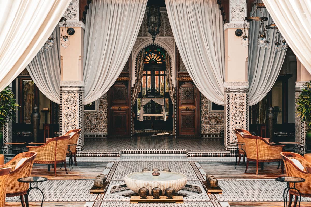 ลานภายในของเรียดโมร็อกโกที่หรูหรา พร้อมน้ำพุกระเบื้อง เก้าอี้ไม้ เสาประดับประดา และประตูโค้งที่กรอบด้วยผ้าม่านสีขาวพลิ้วไหวในเมดินาของมาร์ราเกช