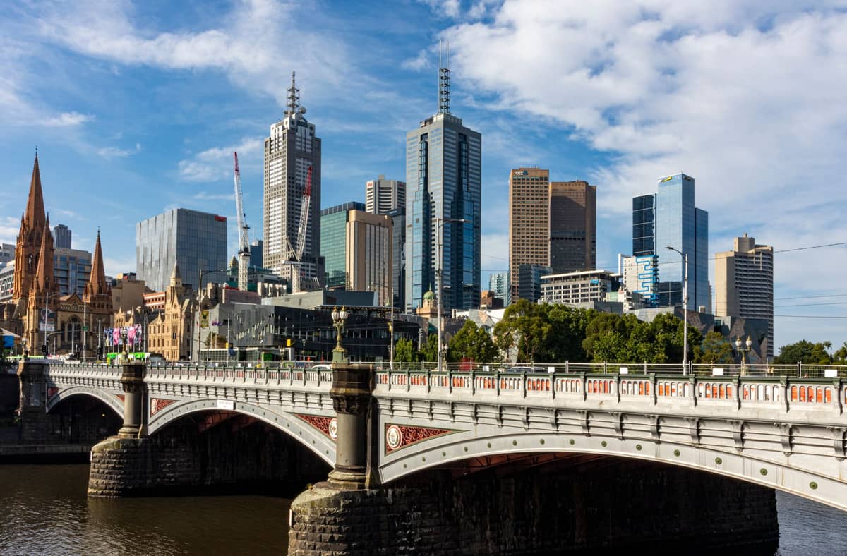 Vista diurna del centro de Melbourne con el Princes Bridge en primer plano, la Catedral de San Pablo, rascacielos modernos y el río Yarra bajo un cielo parcialmente nublado.