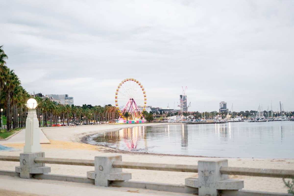 Der St Kilda Beach an einem bewölkten Tag mit der Uferpromenade, Palmen und dem ikonischen Riesenrad im Hintergrund.