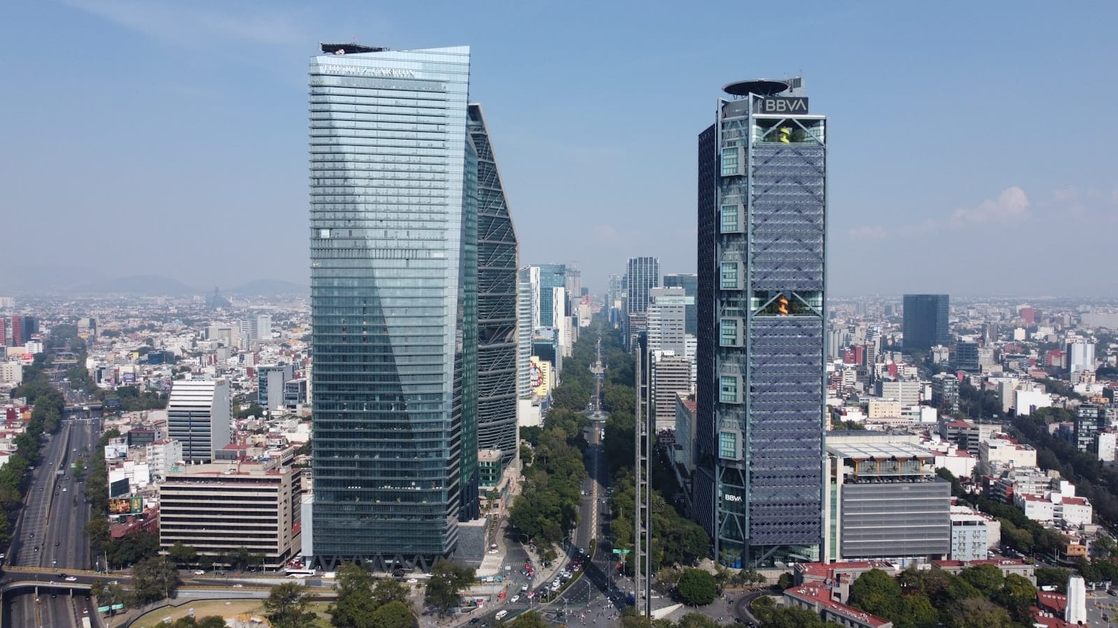 Vista aérea do Paseo de la Reforma na Cidade do México, com arranha-céus modernos, avenida arborizada e bairros se espalhando sob um céu azul.