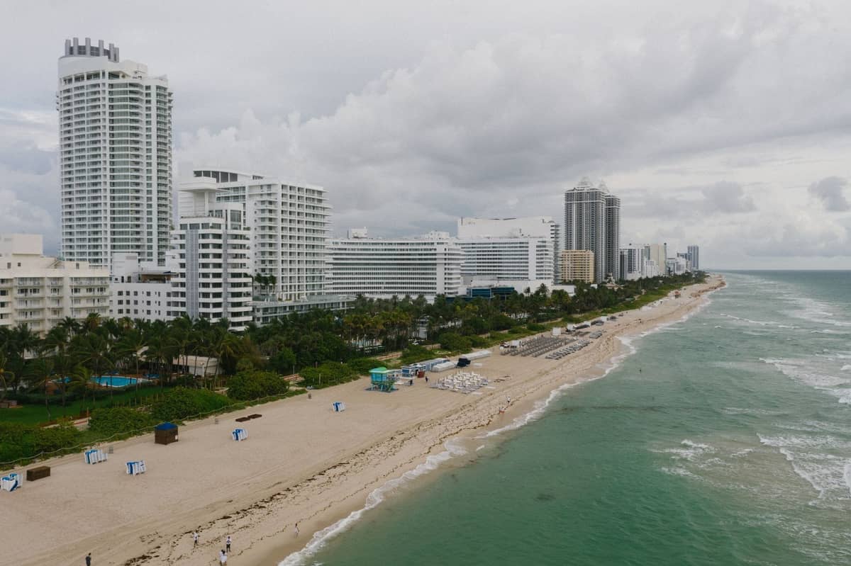 High-rise hotels and condos line a wide, sandy stretch of Miami Beach with few crowds and the ocean to the right under a cloudy sky.