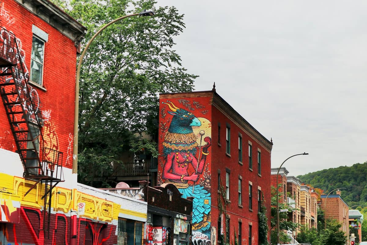 Rote Backsteingebäude mit auffälliger Wandmalerei, schmiedeeisernen Feuertreppen und einer baumgesäumten Straße, typisch für das Plateau-Mont-Royal-Viertel in Montreal.