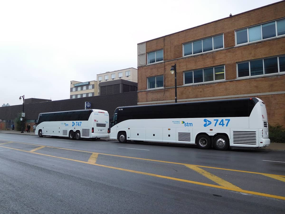 Zwei STM 747 Flughafenshuttlebusse, die an einer Straße in Montreal geparkt sind, mit Gebäuden und einem Metro-Schild im Hintergrund.
