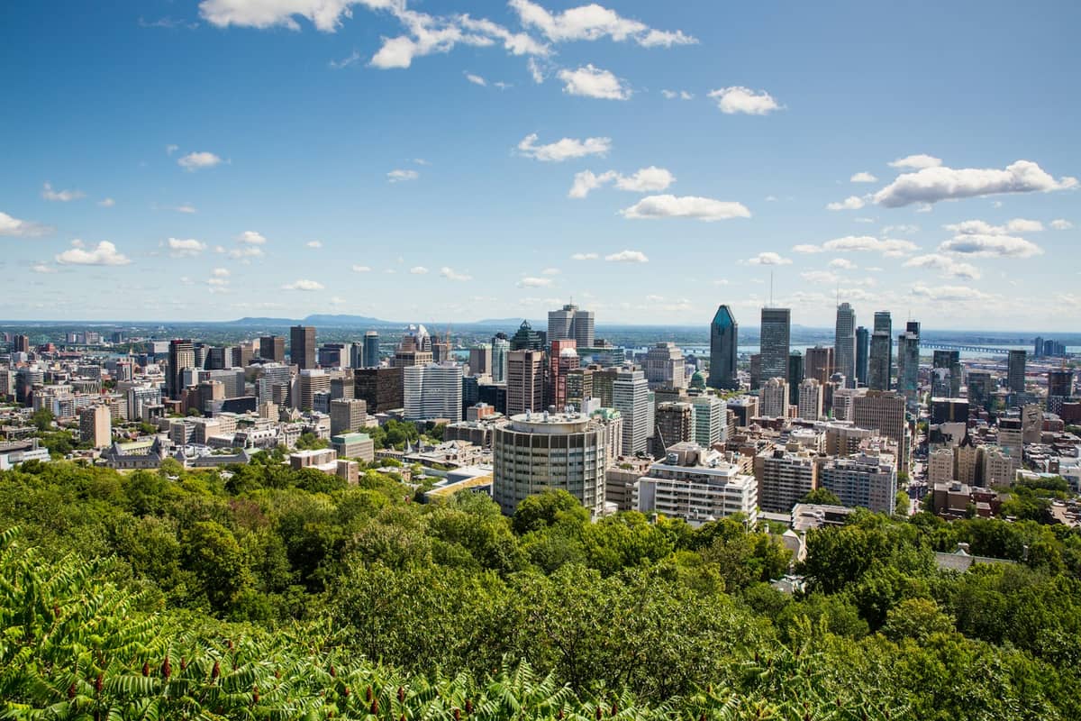 Weiter Tagesblick auf die Skyline der Innenstadt von Montreal mit modernen Hochhäusern und Grünflächen im Vordergrund unter einem blauen Himmel mit Wolken.