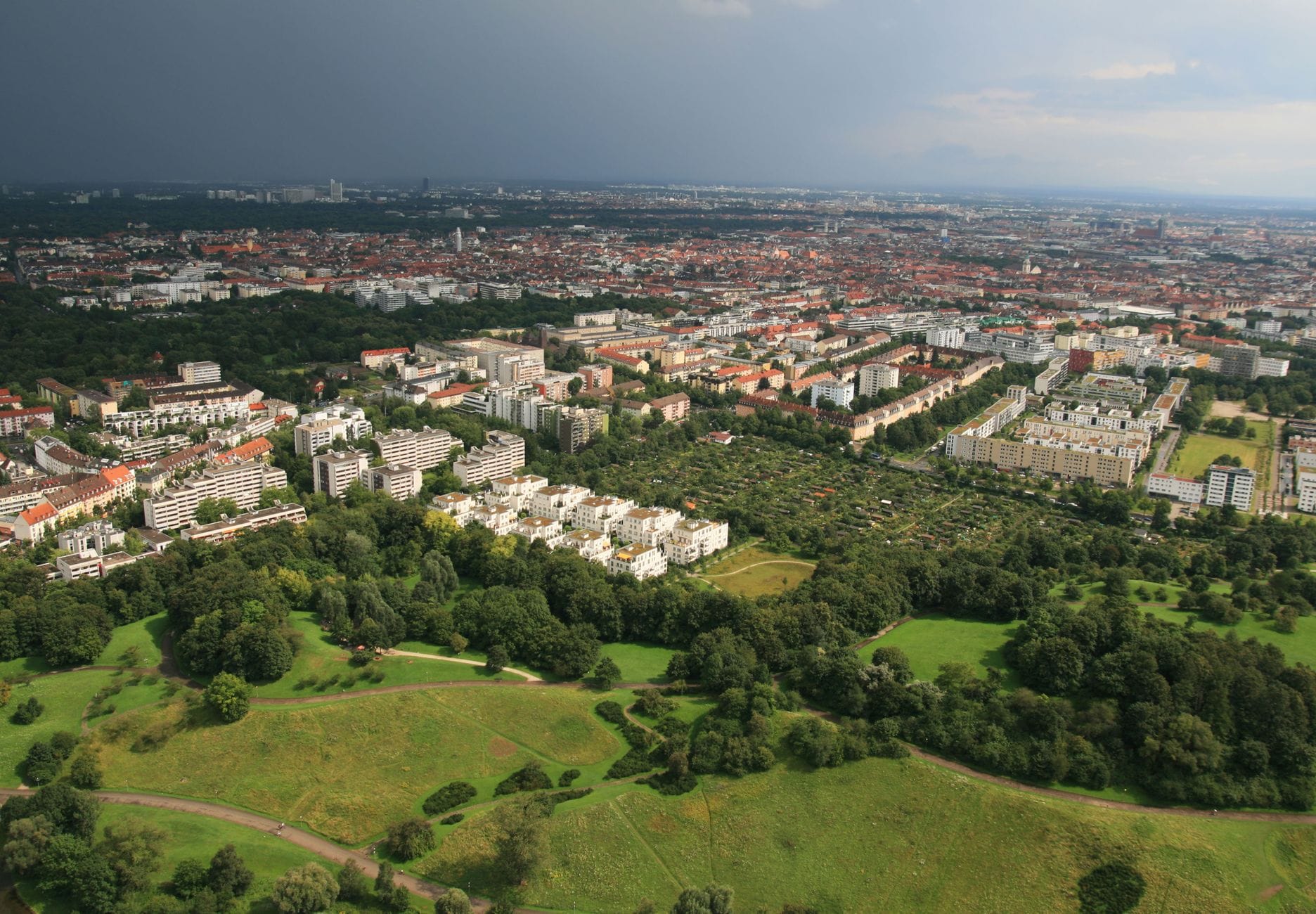 วิวพาโนรามาทางอากาศเหนือใจกลางเมือง Munich ที่แสดงให้เห็นย่านที่อยู่อาศัยหนาแน่น สวนสีเขียว และเส้นขอบฟ้าของเมืองในระยะไกล