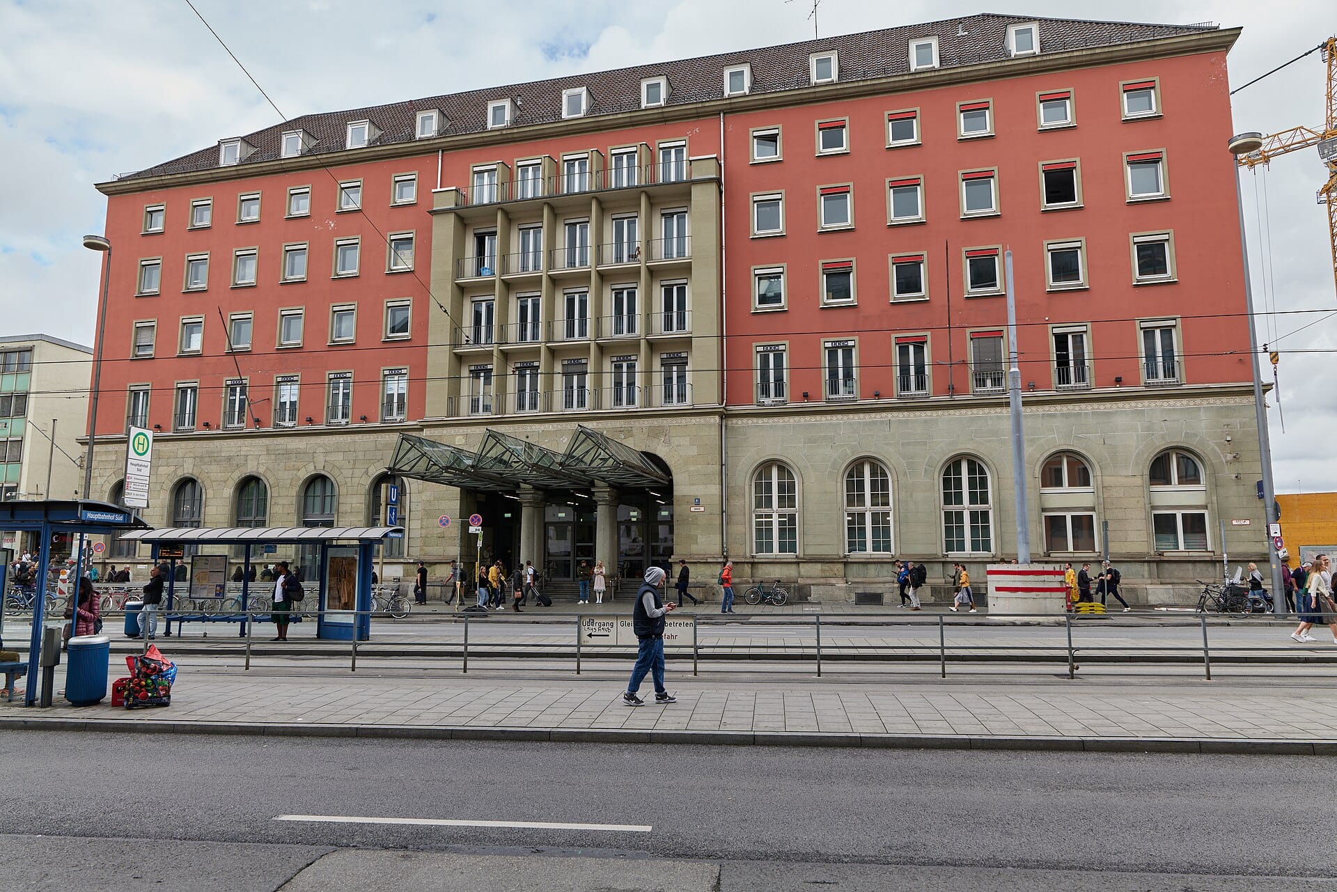 วิวถนนบริเวณทางเข้าหลักของ Munich Hauptbahnhof แสดงให้เห็นด้านหน้าอาคาร ป้ายหยุดรถราง และผู้คนที่เดินอยู่ใกล้เคียง
