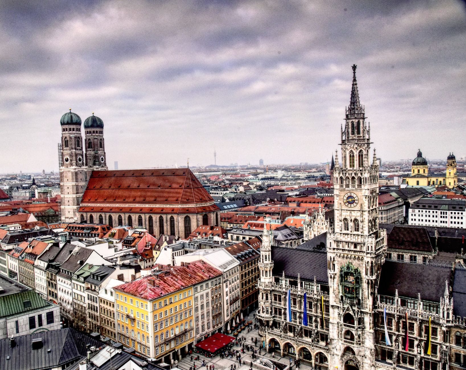 ภาพมุมสูงของมิวนิก เห็นย่าน Marienplatz กับศาลาว่าการใหม่และโบสถ์ Frauenkirche โดดเด่นท่ามกลางหลังคาแดงในเมืองใต้ท้องฟ้าครึ้ม