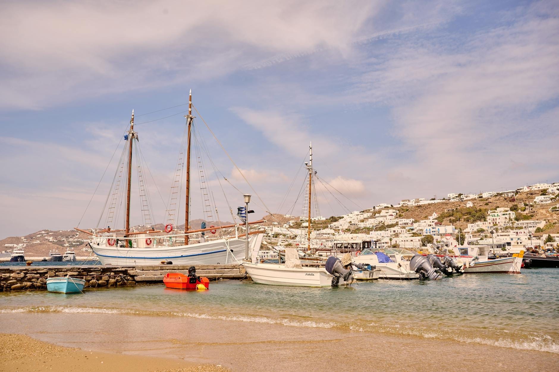 Barcos anclados en una bahía tranquila y poco profunda cerca de una playa de arena, con edificios encalados en una colina al fondo bajo un cielo soleado.