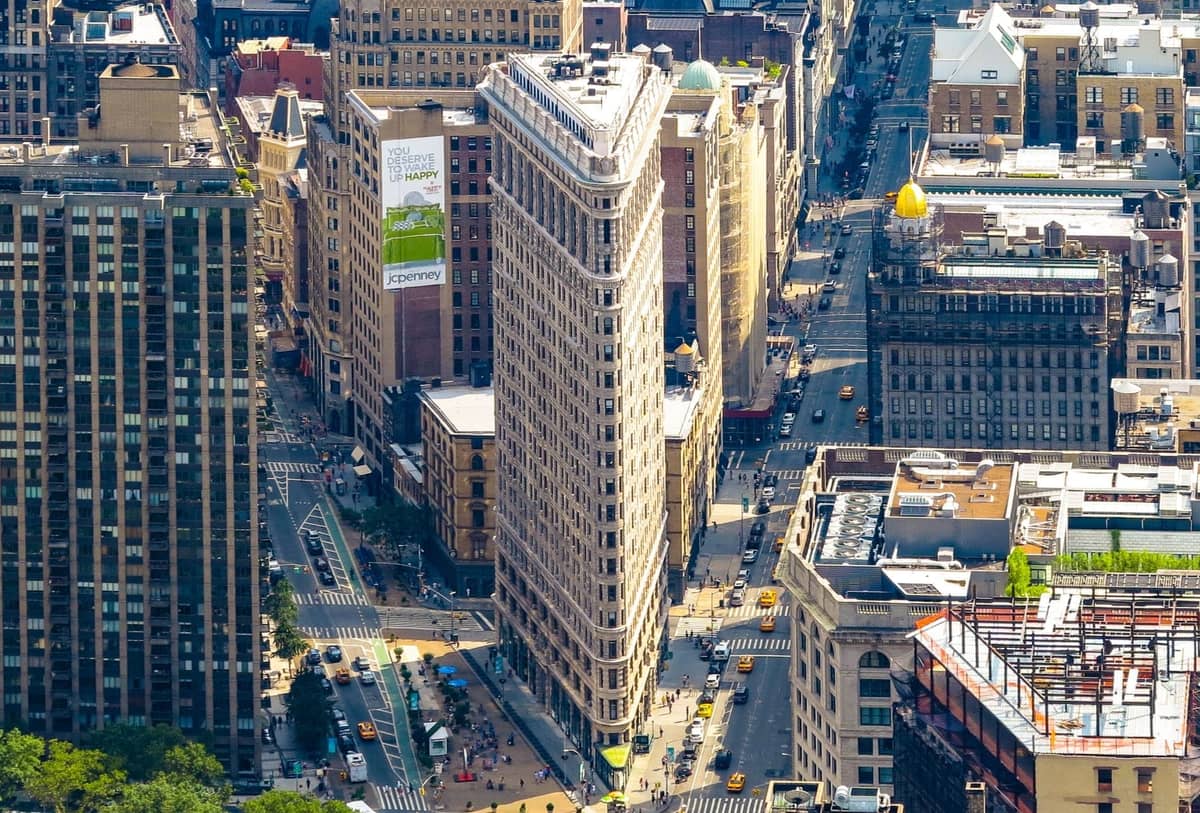 Luftaufnahme des Flatiron Building und der umliegenden Straßen Manhattans bei hellem Tageslicht, die die ikonische dreieckige Architektur und das Stadtleben zeigt.
