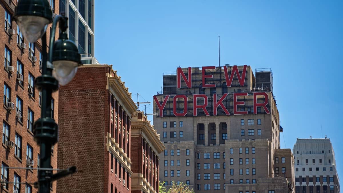 Ikonisches Gebäude des Hotel New Yorker mit großem rotem Schriftzug, umgeben von der Architektur des Midtown Manhattan an einem klaren Tag.