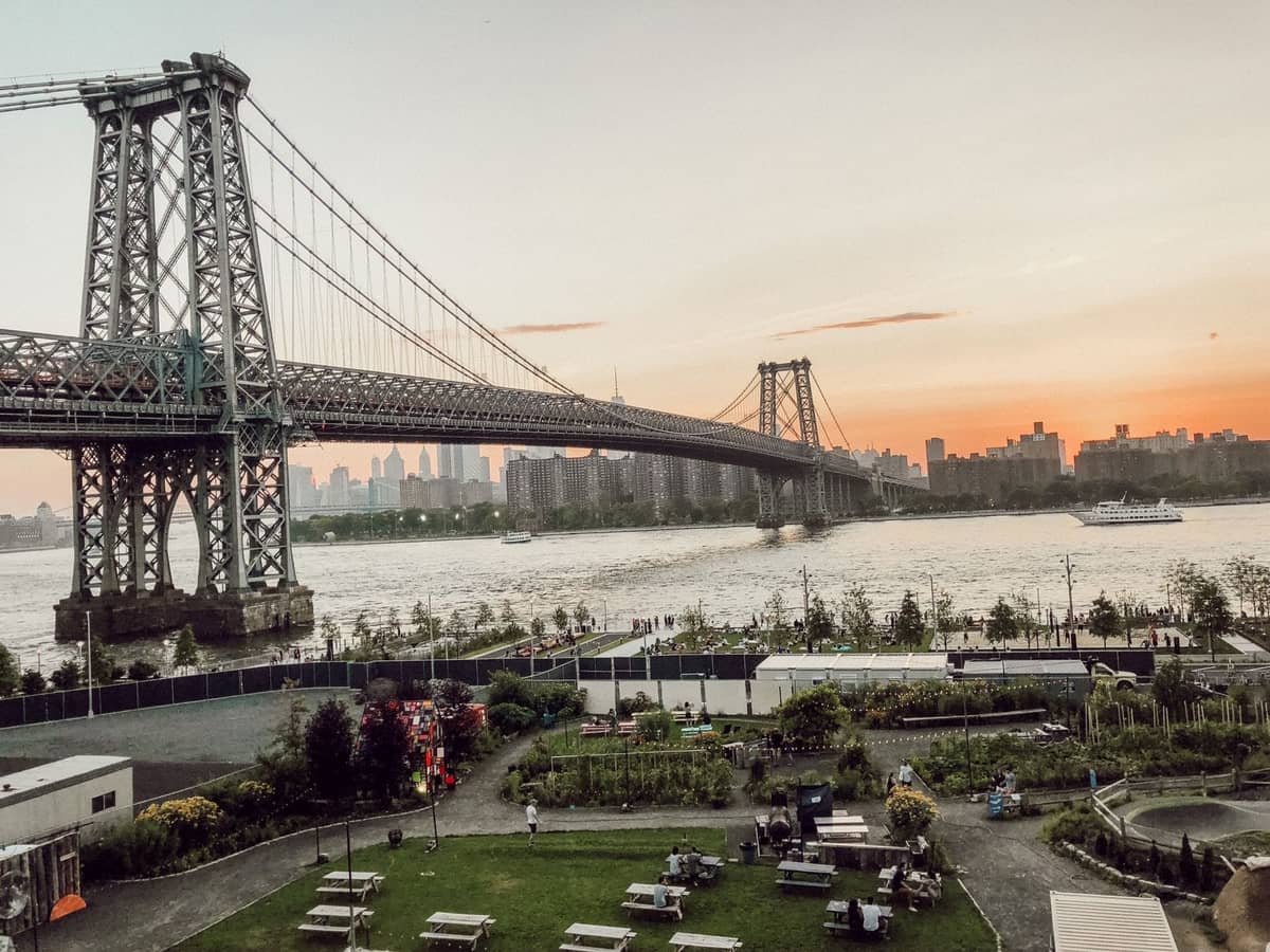 Blick auf die Williamsburg Bridge, die sich über den East River erstreckt, mit der Skyline im Hintergrund und Menschen, die einen grünen Uferpark in Brooklyn genießen.