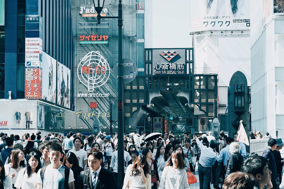 Crowded entrance to Shinsaibashi-suji shopping street in Osaka with many pedestrians, colorful storefronts, and cityscape in daylight.