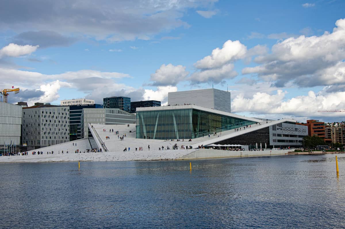 วิวกว้างของ Oslo Opera House โดยมีน้ำอยู่เบื้องหน้าและผู้คนเดินอยู่บนหลังคาสีขาวที่ลาดเอียง