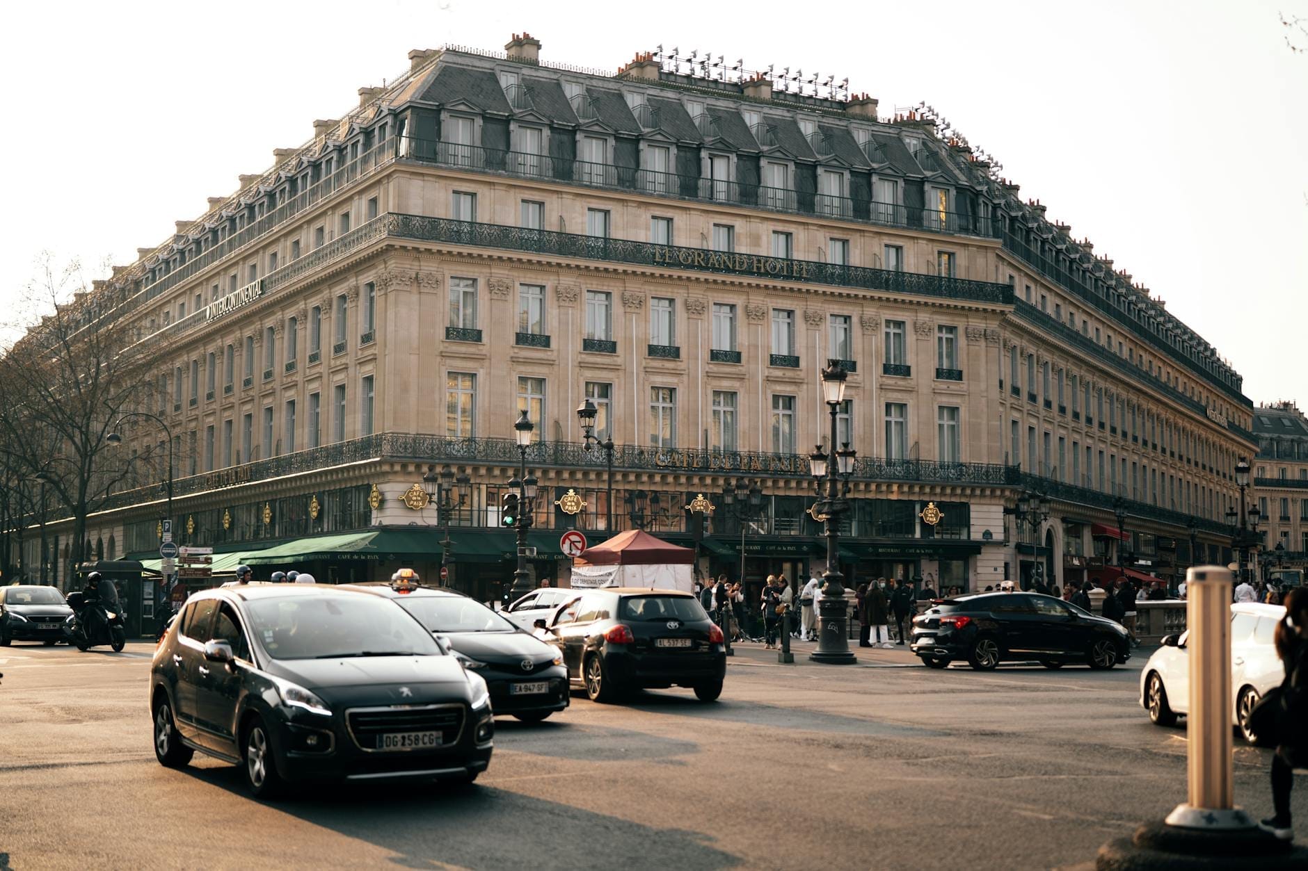 Ampio boulevard parigino con i classici palazzi haussmanniani, auto in movimento, passanti e lampioni eleganti in una giornata di sole, che evoca l'atmosfera vivace dei quartieri di Parigi.