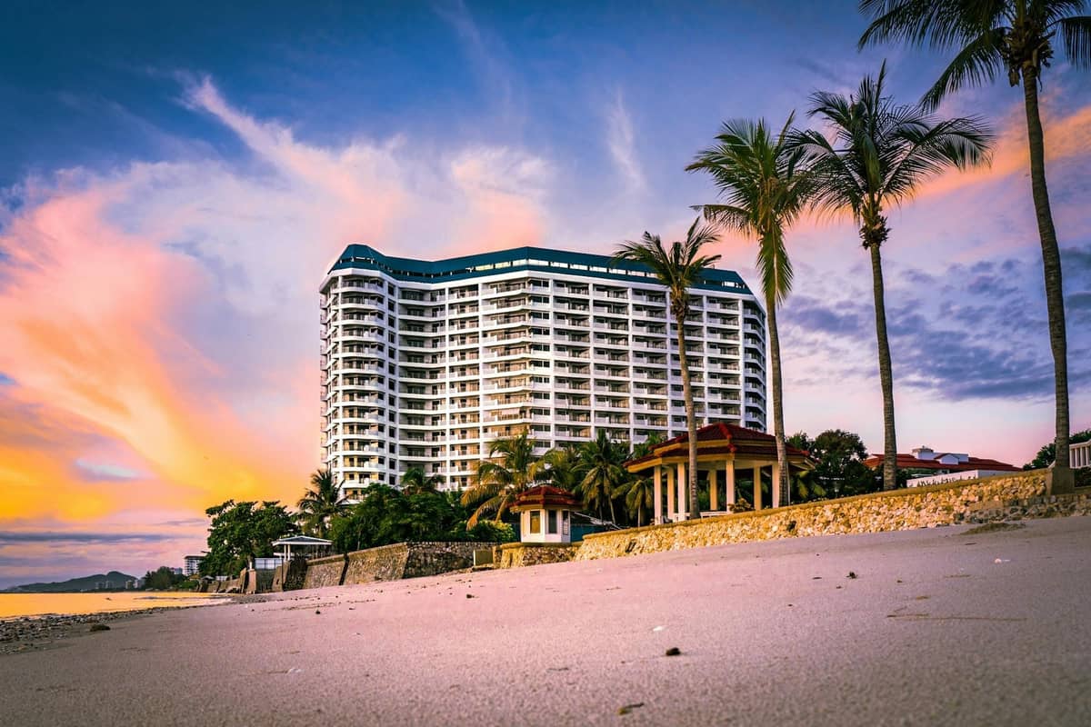 Modern beachfront condominium building at sunset with palm trees and a clean, quiet sandy beach in Pattaya, Thailand.