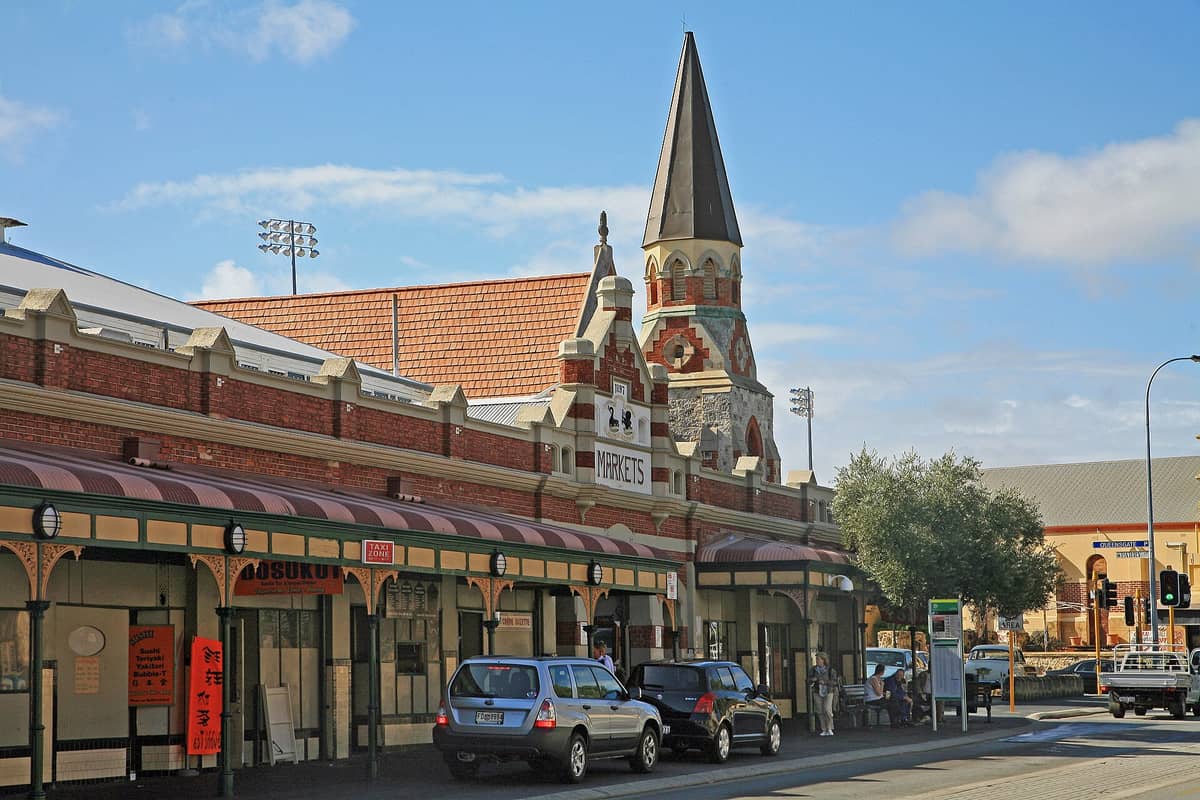 Budynek Fremantle Markets z architekturą wiktoriańską i spiczastą wieżą, zaparkowane przed nim samochody oraz tętniąca życiem ulica w Fremantle, Australia Zachodnia.