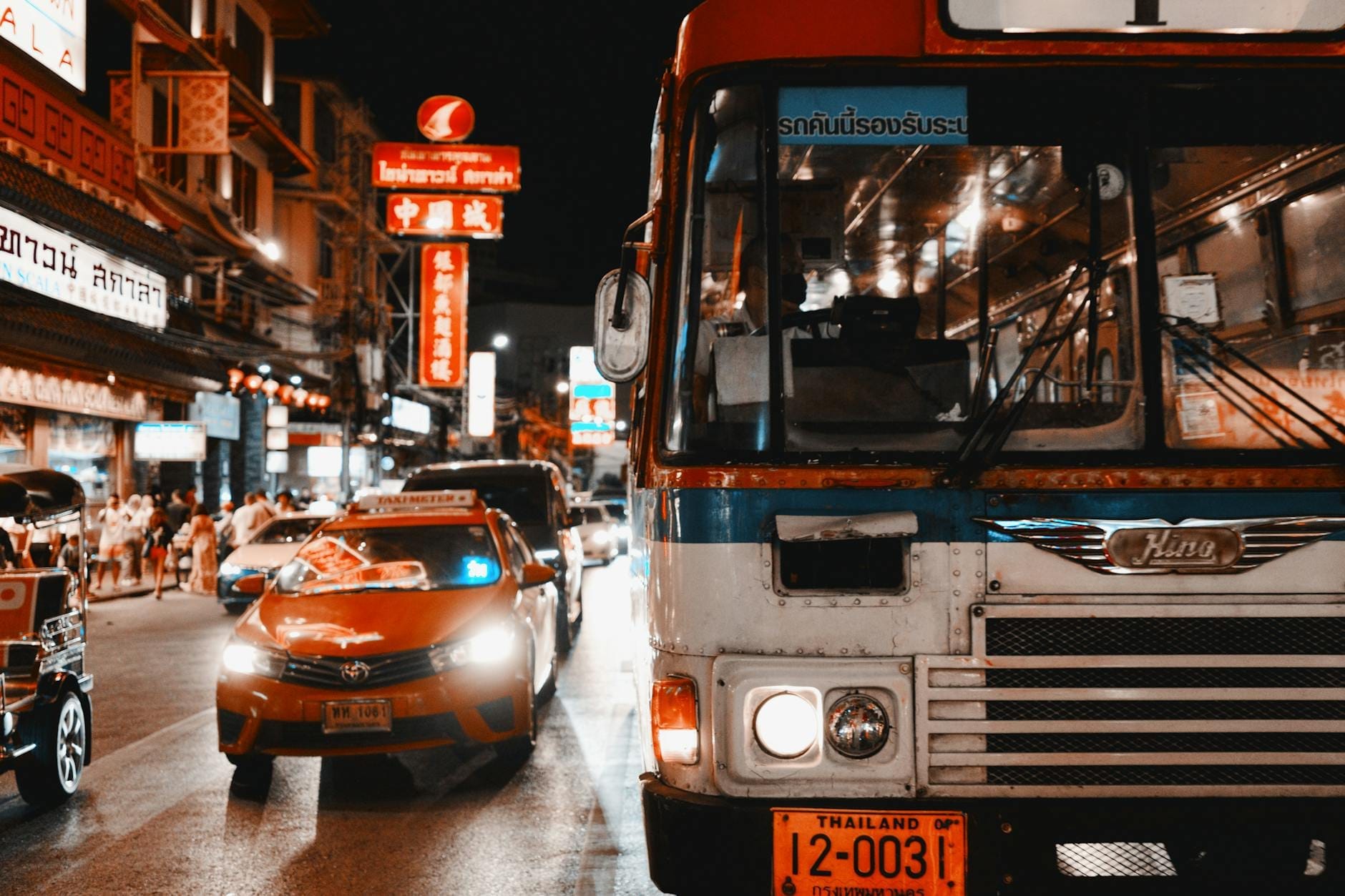 Night street scene in Thailand with a taxi, tuk tuk, and bus amid neon-lit signs, capturing typical local transport.