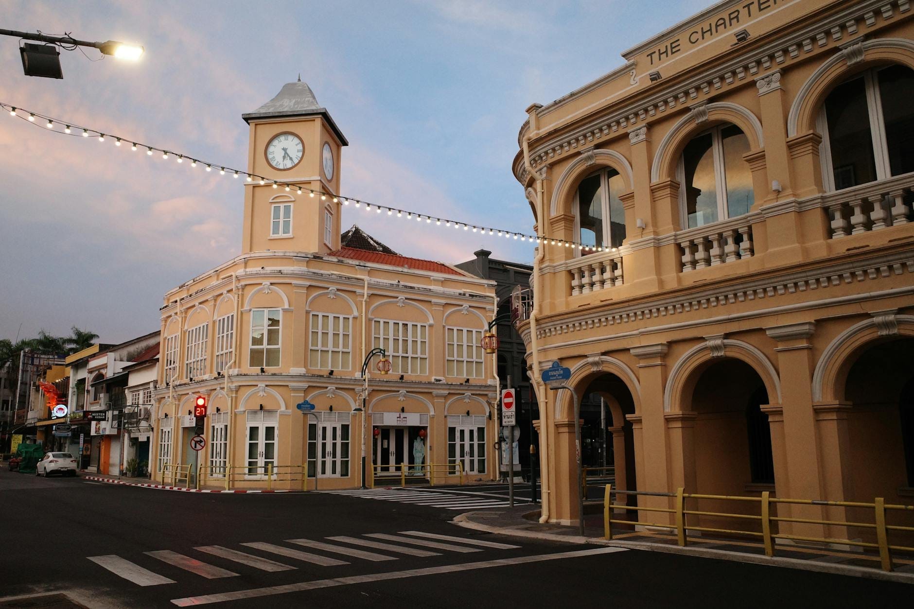 Quiet street corner in Phuket Old Town at dusk, featuring colonial architecture, a yellow clocktower building, and arched balconies.