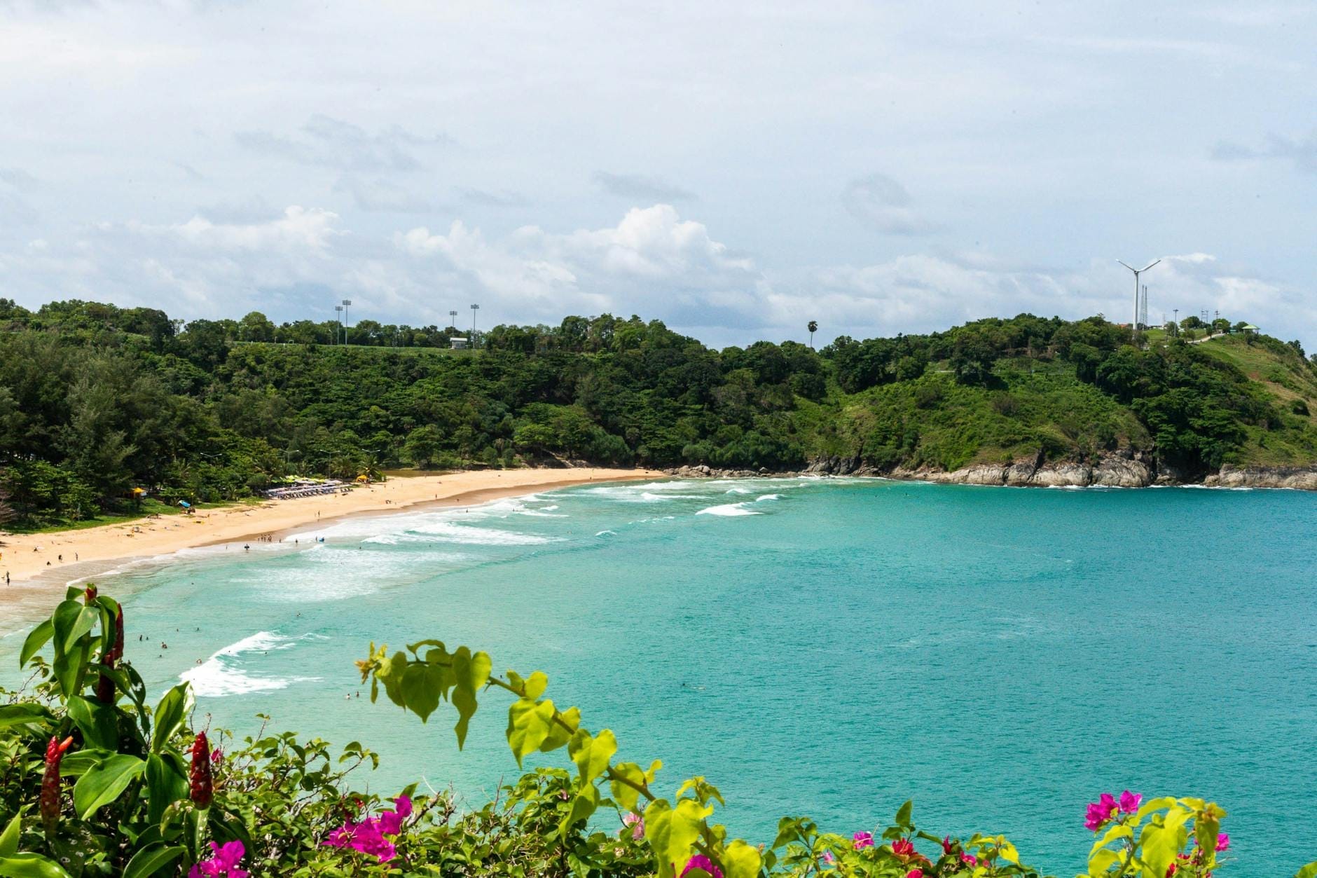 Tropical Phuket beach with turquoise waters, golden sand, lush green hills, and vibrant flowers under a partly cloudy sky.