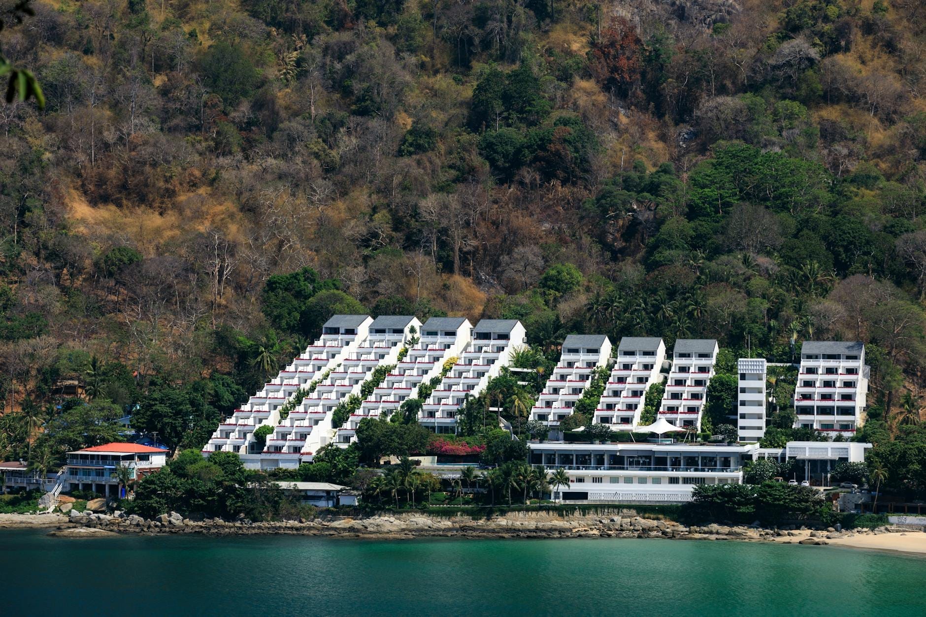 Upscale beachfront resort complex set against a forested Phuket hillside, fronted by turquoise water, embodying the exclusive feel of Kamala, Surin, and Bang Tao.