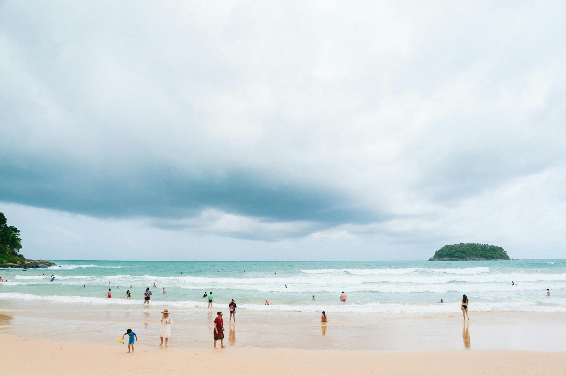 Wide sandy beach with families and children playing in the water on a cloudy day, small green island visible offshore.