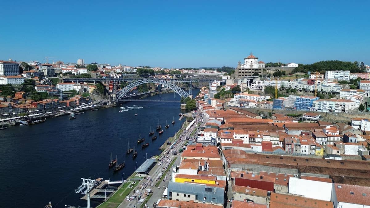 Luftaufnahme der Uferpromenade von Vila Nova de Gaia, des Flusses Douro, der Brücke Dom Luís I und der Skyline von Porto unter einem klaren blauen Himmel.